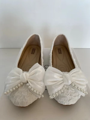 Vita ballerinaskor med rosett och pärlor - Snygga vita ballerinaskor från CUCCOO med stora rosetter framtill dekorerade med pärlor. Skorna har en texturerad yta och rund tå. Perfekta för dig som vill ha en söt och elegant look till din outfit.
