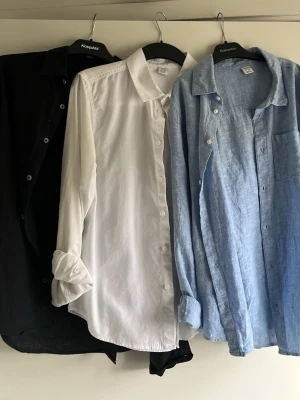 3-pack skjortor  - Säljer tre klassiska skjortor i svart, vit och ljusblå. Alla har lång ärm, bröstficka och knappar framtill. Perfekta för en clean och stilren look. Materialet känns som bomull eller linne, vilket ger en avslappnad vibe.