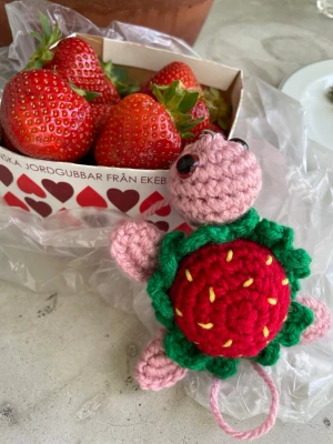 Virkad sköldpadda med jordgubbsskal - Söt handvirkad sköldpadda i rosa garn med ett skal som ser ut som en jordgubbe, dekorerat med gröna blad och gula detaljer. Perfekt som accessoar till väskan eller nyckelringen. Unik och färgglad detalj för dig som gillar gulliga saker.