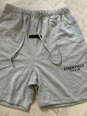 Grå Essentials Fear of God shorts - Snygga grå shorts från Essentials Fear of God med snörning i midjan och tryckt logga på benet. Tillverkade i mjuk bomull för maximal komfort och avslappnad stil. Perfekta för sommardagar eller chill hemma. Klassisk passform och enkel design.