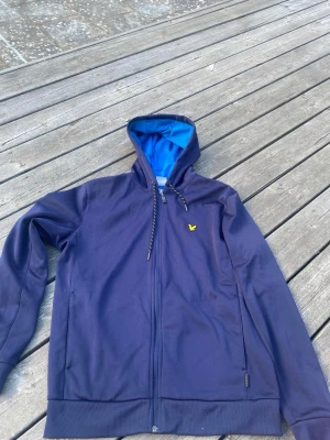Marinblå hoodie från Lyle & Scott - Snygg marinblå hoodie med dragkedja från Lyle & Scott. Har en gul broderad fågel på bröstet.