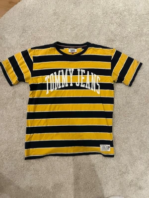 Randig T-shirt från Tommy Jeans - Snygg randig T-shirt från Tommy Jeans i svart och gul med vita detaljer. T-shirten har korta ärmar, rund halsringning och ett stort tryck med 'TOMMY JEANS' på bröstet. Tillverkad i mjuk bomull för skön känsla. Perfekt för en avslappnad och trendig look.