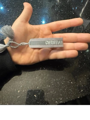 Grått Christian Dior J'Adior armband - Snyggt grått armband från Christian Dior med texten 'J'ADIOR' och 'CHRISTIAN' invävt. Armbandet är gjort i tyg och har dekorativa tofsar i ändarna. Perfekt accessoar för att lyfta din outfit med en exklusiv touch.
