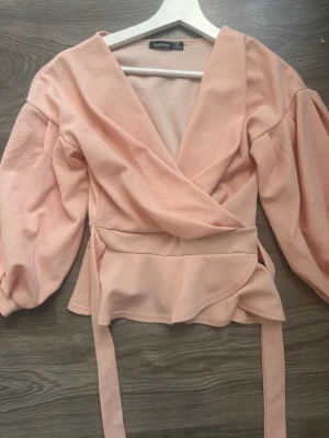 Ljusrosa omlottblus från Boohoo - Trendig ljusrosa omlottblus från Boohoo med korta puffärmar och knytband i midjan. Blusen har en djup v-ringning och är tillverkad i ett strukturerat, lätt tyg som ger en snygg och modern look. Storlek 36 men passar även 34 och kanske 32 (PRISET GÅR ATT DISKUTERA KAN SÄNKA HUR MYCKET SOM HELST) har kortare ärm
