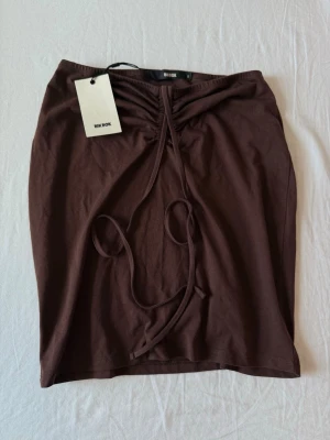 Brun kjol med snörning från Bik Bok - Snygg brun kortkjol från Bik Bok med trendig snörning framtill. Storlek S. Kjolen har en enkel, rak passform och är tillverkad i mjukt bomullsmaterial. Perfekt att styla med en crop top eller oversized tröja för en cool look. Helt ny. Aldrig använt kjolen. Prislapp kvar. Orginalpris 199kr. 