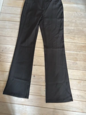 Svarta kostymbyxor från Outfitbook - Säljer ett par svarta kostymbyxor från Outfitbook med utsvängda ben och hög midja. Byxorna har en klassisk look och är tillverkade i ett slätt tyg som ger en snygg siluett. Perfekta för dig som gillar stilrena och trendiga plagg.