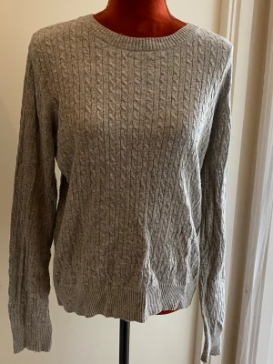 Grå kabelstickad tröja M - Tröja med rund halsringning och långa ärmar. Tröjan har ett flätstickat mönster över hela plagget, vilket ger en klassisk och texturerad look. Muddar finns vid ärmslut och nederkant.Längd: 62 cm Bystvidd: 55 cm 50% bomull 25% polyamid 25% viskos  Inga bilder på mig! Katter finns i hemmet!