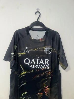 PSG x Jordan svart matchtröja - Snygg svart PSG matchtröja i samarbete med Jordan, med marmorerat mönster i gult och rött. Tröjan har korta ärmar, rund hals och tryckta loggor för både PSG och Jordan samt Qatar Airways som sponsor. Tillverkad i lätt och ventilerande funktionsmaterial.