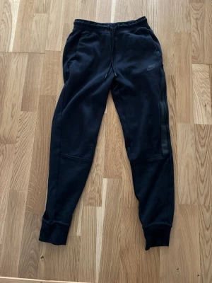 Svarta Nike mjukisbyxor med dragkedja - Säljer ett par svarta mjukisbyxor från Nike med smal passform och resår i midjan. Byxorna har dragsko, ribbade muddar vid bensluten och en snygg dragkedjeficka på sidan. Perfekta för chill eller träning.