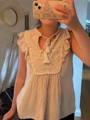 Beige blus med broderi och volanger - Söt beige blus med broderade detaljer framtill, volangkantade ärmhål och knytband med tofsar vid halsen. Den är enbart testad men aldrig använd. Passar 34/36 (Xs/S)