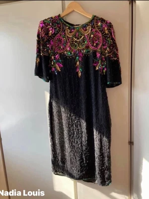 Svart klänning med paljetter Nadia Louis - Unik svart klänning från Nadia Louis med färgglada paljetter och broderier i guld, rosa, grön och blå. Klänningen har kort ärm, rund halsringning och vackra detaljer upptill. Perfekt för dig som vill sticka ut med en glamorös look.passar s-m kanske även L