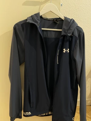 Mörkblå vindjacka från Under Armour - Snygg mörkblå vindjacka från Under Armour med huva och dragkedja framtill. Jackan har vita logodetaljer och elastiska muddar vid ärmsluten. Tillverkad i lätt och smidigt material som passar perfekt för blåsiga dagar.