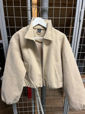 Beige bomberjacka med dragkedja  - Stilren beige bomberjacka med dragkedja framtill och bred krage. Jackan har en loose passform, två sidofickor och är tillverkad i ett mjukt material som känns som ull eller fleece.