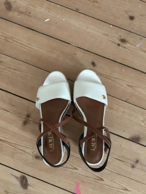 Vita kilklackade sandaler Ralph Lauren - Säljer ett par snygga sandaler från Lauren Ralph Lauren med vit ovandel, bruna remmar och svart hälparti. Kilklack i repmaterial ger en somrig känsla. Perfekta för dig som vill ha både stil och höjd. Märkeslogga i guld på ovandelen.