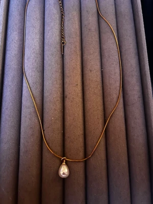Guldigt halsband med pärlhänge - Snyggt halsband i guldfärgad metall med ett droppformat pärlhänge. Kedjan är tunn och elegant med justerbar längd tack vare förlängningskedjan. Perfekt för dig som gillar stilrena accessoarer med en touch av klassisk vibe.