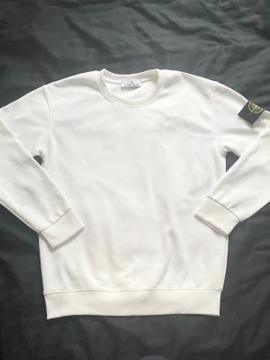 Vit sweatshirt från Stone Island - Säljer en clean vit sweatshirt från Stone Island med klassisk rund hals och ribbade muddar. Tröjan har den ikoniska loggan på vänster ärm och är tillverkad i mjukt bomullsmaterial. Perfekt för dig som gillar stilrena och trendiga plagg.