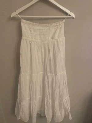 Vit långkjol från Gina Tricot - Luftig och somrig vit långkjol från Gina Tricot med smock i midjan och volangdetaljer. Kjolen har en avslappnad passform och är tillverkad i ett mjukt bomullstyg som passar perfekt till varma dagar. Aldrig använd!
