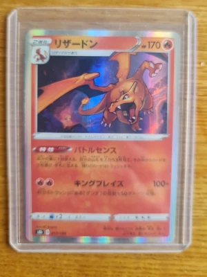 Japansk Charizard Pokémonkort - Säljer ett japanskt Charizard Pokémonkort med holografisk yta. Kortet har starka färger i orange, rött och blått och ligger i ett genomskinligt plastfodral. Perfekt för samlare eller Pokémonfans som vill utöka sin samling.