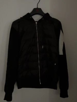 Svart pufferjacka från Moncler - Snygg svart pufferjacka från Moncler med huva och dragkedja framtill. Jackan har quiltad design, ribbade muddar och ett vit M på ena ärmen. Även NFC funkar! Perfekt för sommarkvällar och dig som vill gå runt med en schysst look! (PRISET GÅR ATT DISKUTERAS) 🫱🏽‍🫲🏼