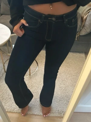 Mörkblå bootcut jeans med kontrastsömmar - Snygga mörkblå bootcut jeans med vita kontrastsömmar och klassisk femficksdesign. Jeansen har hög midja och utsvängda ben, vilket ger en trendig siluett. Perfekta att matcha med crop tops eller hoodies för en cool look.