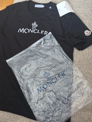 Svart Moncler t-shirt med logga - Snygg svart t-shirt från Moncler med vit logotyp på bröstet och ett broderat Moncler-märke på ärmen. Klassisk rund halsringning och korta ärmar. Tillverkad i mjuk bomull för en skön känsla. Perfekt för dig som gillar stilrena plagg med exklusiv känsla.