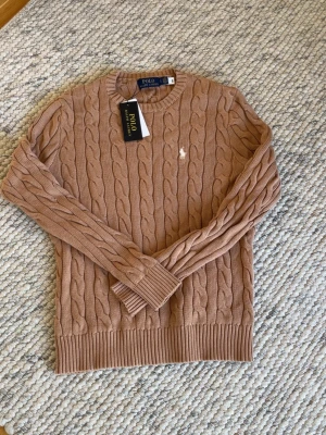 Beige kabelstickad tröja Polo Ralph Lauren - Kabelstickad tröja i beige från Polo Ralph Lauren med rund halsringning och ribbade muddar. Tröjan har den klassiska broderade loggan på bröstet och är tillverkad i mjukt material. Perfekt för dig som gillar stilrena och tidlösa plagg.
