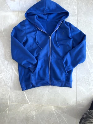 Blå hoodie från Polo Ralph Lauren - Blå hoodie med dragkedja från Polo Ralph Lauren. Klassisk design med huva och dragsnören, två fickor framtill och broderad logga på bröstet. Tillverkad i mjuk bomullsblandning som känns skön mot huden. Perfekt för en avslappnad och stilren look.