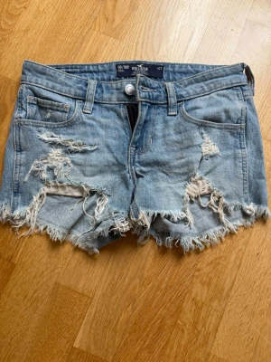 Ljusa slitna jeansshorts Hollister - Säljer ett par ljusblå jeansshorts från Hollister med slitna detaljer och fransig kant. Klassisk femficksmodell med knapp och dragkedja framtill. Perfekta för sommaren och ger en avslappnad vibe. midjemåttet är cirka 38 cm