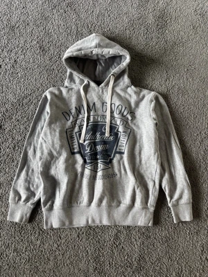 Hoodie - Snygg grå hoodie från Denim Goods med stort svart tryck på bröstet där det står 'Authentic Denim'. Hoodien har huva med vita dragsnören och ribbade muddar vid ärmar och nederkant. Perfekt för en avslappnad streetwear-look.