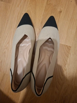 Beige ballerinaskor med svart tå - Snygga ballerinaskor i beige med svart spetsig tå och svart kant. Skorna har en stickad ovandel och fyrkantig klack i svart och beige. Perfekta för dig som gillar en modern och stilren look.