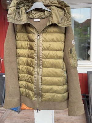 Olivgrön cardiganjacka från Moncler - Snygg olivgrön cardiganjacka från Moncler med quiltad front och huva i glansigt material. Ärmar och rygg i mjuk, ribbstickad ullmix. Dragkedja framtill och på ärmen samt Moncler-logga på armen. Perfekt för lager på lager och streetwear-vibe.