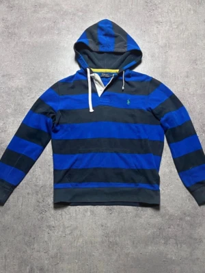 Vintage Polo Ralph Lauren Hoddie - Snygg hoodie från Polo Ralph Lauren med breda blå och svarta ränder. Klassisk huva med vita snören och känguruficka framtill. Liten grön logga broderad på bröstet. Tillverkad i mjuk bomull för skön känsla och avslappnad stil.