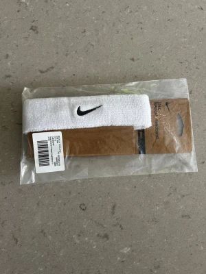 Vit Nike pannband med broderad logga - Snyggt vitt pannband från Nike med den klassiska svarta swoosh-loggan broderad framtill. Tillverkat i mjukt och stretchigt material som passar perfekt för träning eller sport. Enkel och stilren design som håller håret på plats.