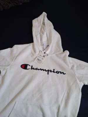Vit Champion hoodie med logga - Snygg vit hoodie från Champion med stor broderad logga i svart och rött på bröstet. Klassisk känguruficka framtill och stor huva. Tillverkad i mjukt bomullsmaterial som är skönt att bära. Perfekt för en avslappnad och sportig stil.