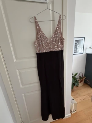 Svart långklänning med paljetter - Elegant långklänning med svart kjol och ljus, v-ringad överdel dekorerad med paljetter. Klänningen har smala axelband och djup urringning både fram och bak. Perfekt för festliga tillfällen där du vill sticka ut.