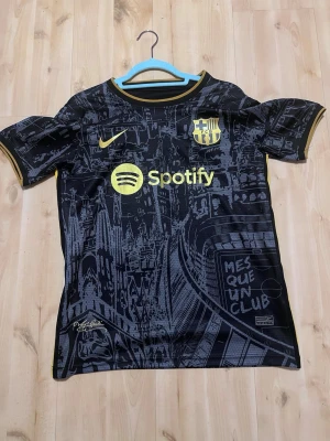 Barcelona matchtröja svart/gul Nike - Snygg svart Barcelona matchtröja med gula detaljer och tryck, inklusive Spotify-logga och klubbmärke på bröstet. Nummer 10 på ryggen och UNHCR-logga. Unik grafisk design med stadsmotiv och texten 'MES QUE UN CLUB'. Perfekt för fotbollsfans!