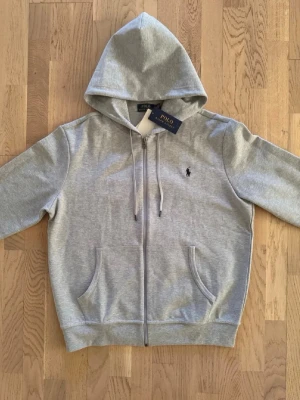 Grå hoodie från Polo Ralph Lauren - Snygg grå hoodie från Polo Ralph Lauren med dragkedja, huva med dragsko och två fickor framtill. Klassisk logga broderad på bröstet. Tillverkad i mjuk bomullsblandning som känns skön mot huden. Perfekt för en avslappnad och stilren look.