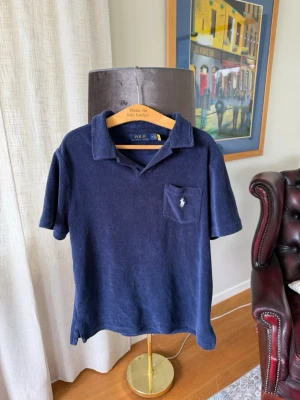 Marinblå frottépiké från Polo Ralph Lauren - Klassisk marinblå pikétröja i mjuk frotté från Polo Ralph Lauren. Tröjan har korta ärmar, krage och en bröstficka med den ikoniska broderade loggan. Perfekt för en avslappnad och stilren look.