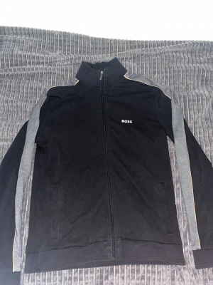 Svart zip hoodie från BOSS - Svart hoodie från BOSS med grå detaljer längs ärmarna och en tunn gul linje. Stängs med dragkedja och har två fickor framtill. Materialet är mjukt och bekvämt, perfekt för en avslappnad look. BOSS-loggan är broderad på bröstet.