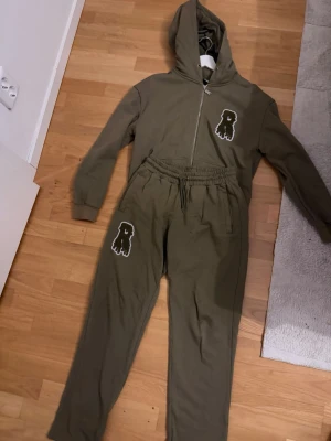 Olivgrön hoodie-set med tryck - Mysigt loungewear-set i olivgrönt med matchande hoodie och byxor. Setet har dragkedja framtill, huva och elastisk midja. Både tröjan och byxorna har ett vitt och svart monster-tryck. Perfekt för chill dagar hemma.