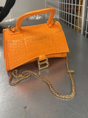 Orange handväska - Snygg orange handväska i krokopräglat konstläder. Väskan har ett guldfärgat B-spänne framtill, matchande guldkedja och ett stadigt handtag. Perfekt statement piece för dig som vill sticka ut med färg och trendig design.