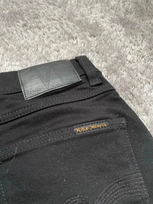 Svarta Lean Dean Dry Nudie Jeans - Säljer ett par helt nya svarta skinny jeans från Nudie Jeans. Klassisk femficksmodell med diskret logga på bakfickan och svart läderpatch i midjan. Jeansen har smal passform och är tillverkade i stretchigt bomullsmaterial för extra komfort.
