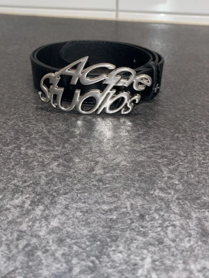 Svart Acne Studios bälte i skinn - Snyggt svart bälte från Acne Studios med ett stort silvrigt metallspänne där märkesnamnet står i cool design. Bältet är gjort i skinn och passar perfekt för att lyfta vilken outfit som helst.