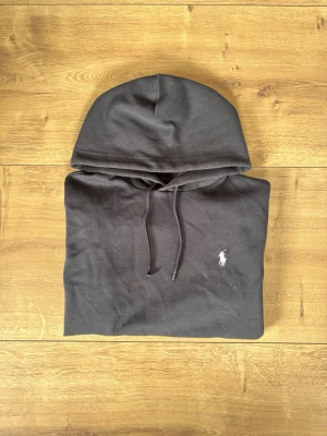 Svart hoodie från Ralph Lauren - Snygg svart hoodie från Ralph Lauren med klassisk huva och dragsnören. På bröstet finns den ikoniska vita broderade loggan med polospelaren. Hoodien är tillverkad i mjukt bomullsmaterial och har en avslappnad passform som passar perfekt till jeans eller joggers.