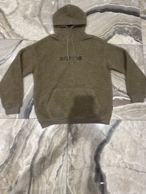 Beige teddyhoodie med japansk text - Mysig beige hoodie i teddy-material med huva och dragsnören. På bröstet finns en broderad japansk text i svart. Hoodien har ribbade muddar vid ärmar och nederkant samt en stor magficka. Perfekt för dig som gillar streetwear och vill ha något riktigt skönt.