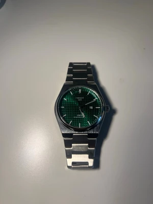 Tissot PRX  - Stilren Tissot PRX 80 Quartz klocka med grön urtavla och datumvisning. Klockan har armband i borstat stål och en modern, kantig form. Perfekt för dig som gillar klassisk design med en twist. skriv bara för mer information.                     Ingen box eller papper tillgår, kommer dock med 3 extra links