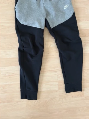 Nike mjukisbyxor svart/grå S - Snygga mjukisbyxor från Nike i svart och grått med dragsko i midjan och ribbade muddar vid benslut. Byxorna har en modern passform och är tillverkade i mjukt bomullsmaterial. Perfekta för chill eller träning. Nike-logga på vänster lår.