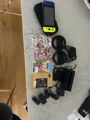 Nintendo Switch med tillbehör och spel - Nintendo Switch med blå och gul Joy-Con, svart dockningsstation, laddare, rattar, handkontroll, fodral och tre spel: Mario Kart 8 Deluxe, Super Mario Party och Crash Team Racing. Perfekt för gaming hemma eller på språng.