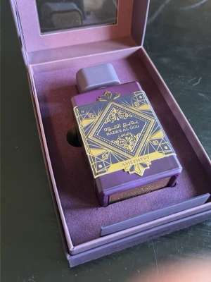 Lattafa Badee Al Oud Amethyst parfym - Lattafa Badee Al Oud Amethyst är en parfym i en lyxig lila flaska med gulddetaljer och orientaliskt mönster. Flaskan har en fyrkantig form och kommer i en matchande lila presentförpackning med fönster. Doften är känd för sina rika och djupa oud-noter.