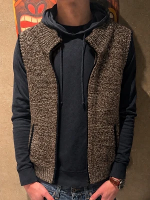 Stickad väst - Säljer en brunmelerad stickad väst med ribbade kanter och hög krage. Perfekt att slänga över en hoodie för en chill och unik mook. Modellen är 185cm